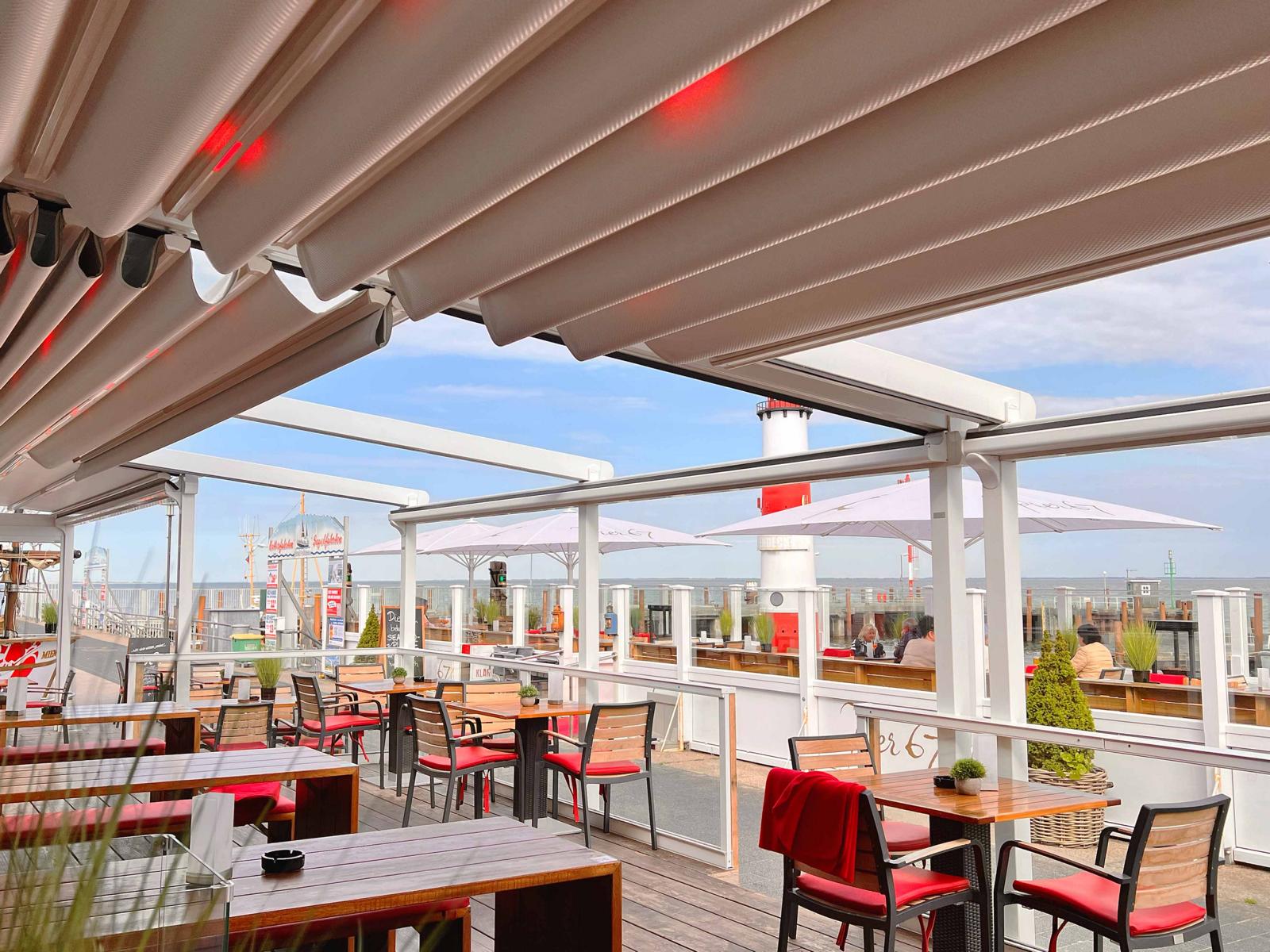 Weißes, halb aufgefahrenes Pergoladach überspannt die Gastronomiefläche vom Pier 51 von Gosch auf Sylt - mit integrierten LED-Strahlern, die mit rotem Licht akzentuieren. Im Hintergrund Blick auf den Leuchtturm und den Hafen.